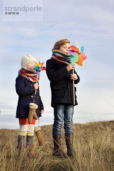 Bruder und Schwester blasen auf Papierwindmühlen an der Küste