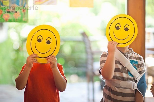Zwei Jungen halten Smiley-Gesichtsmasken im Kindergarten hoch.