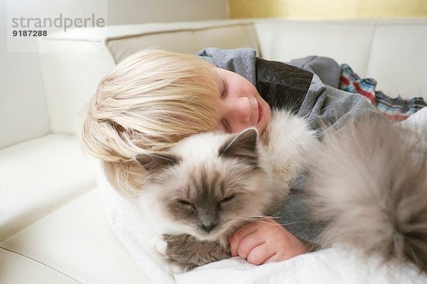 Süßer dreijähriger Junge mit geschlossenen Augen  der Katze auf dem Sofa umarmt