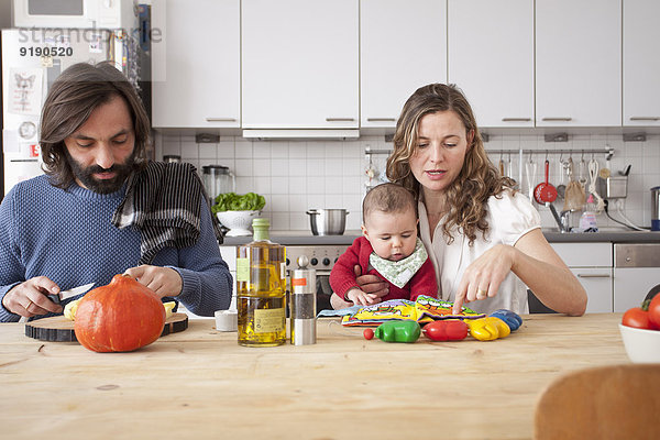 Vater schneidet Gemüse mit der Mutter  die mit dem Mädchen in der Küche spielt.