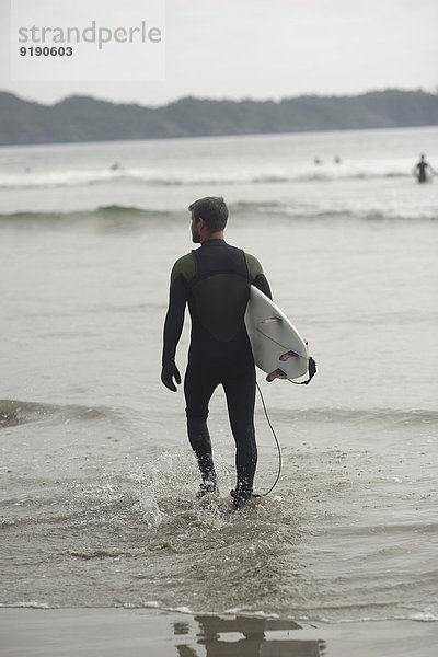 Durchgehende Rückansicht des Mannes mit Surfbrett im Wasser am Strand