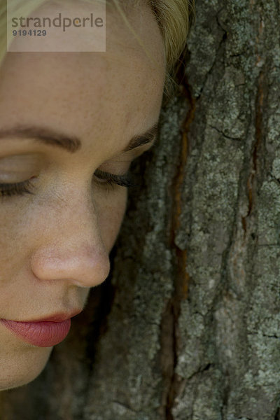 Junge Frau lehnt sich an den Baumstamm und schaut traurig nach unten.