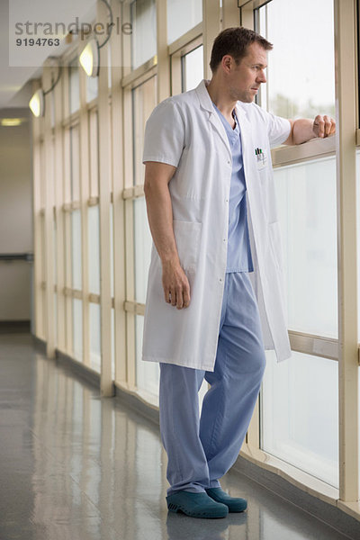 Männlicher Arzt steht im Krankenhaus und schaut aus dem Fenster.