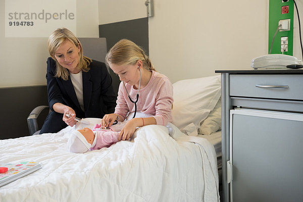Mädchen untersucht eine Puppe mit Stethoskop und ihre Mutter sitzt neben ihr im Krankenhaus.