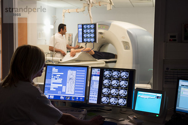Ärztin untersucht Scan am Computer mit Patientin auf MRT-Scanner im Hintergrund