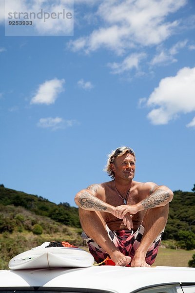 Erwachsener Mann mit Surfbrett auf dem Fahrzeugdach am Strand sitzend