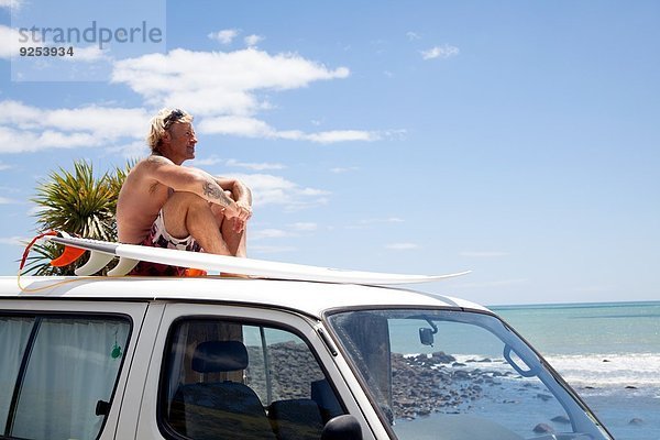 Erwachsener Mann mit Surfbrettbeobachtung vom Fahrzeugdach am Strand aus