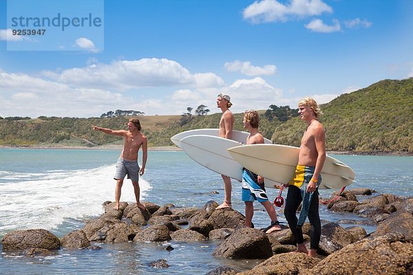 Vier junge Surferfreunde  die von Felsen auf das Meer zeigen.