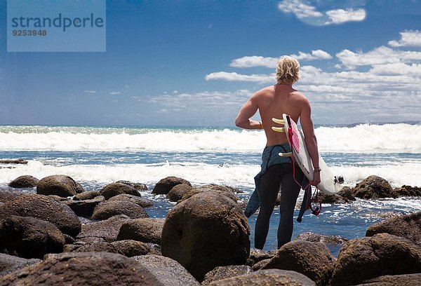 Junge erwachsene männliche Surfer beobachten das Meer von den Strandfelsen aus.
