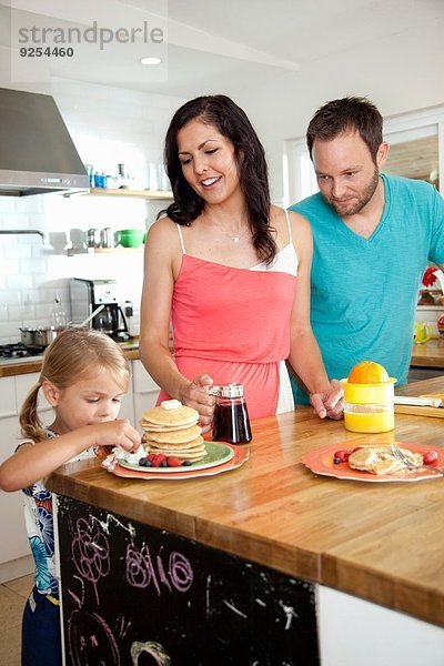 Mittlere erwachsene Eltern beobachten die Tochter  die von Pfannkuchen an der Frühstücksbar versucht wird.