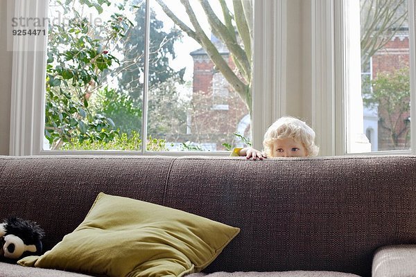 Porträt eines süßen blonden Kleinkindes  das sich hinter dem Sofa versteckt.