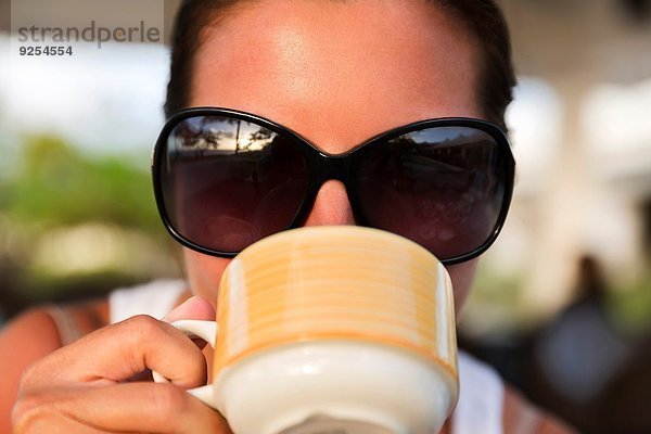 Nahaufnahme einer erwachsenen Frau in einer Sonnenbrille  die aus einer Kaffeetasse trinkt.