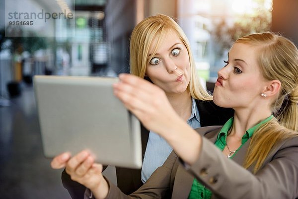 Zwei junge Geschäftsfrauen ziehen Gesichter und nehmen Selfie auf digitalem Tablett im Büro mit.