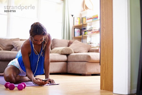 Junge Frau  die auf dem Boden des Wohnzimmers trainiert  während sie auf das Smartphone schaut.