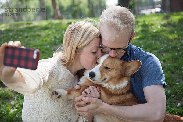 Junges Paar im Park nimmt Selfie während er Corgi Dog küsst.