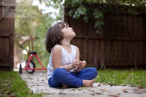 Vierjähriges Mädchen sitzt auf dem Gartenweg und blickt nach oben.