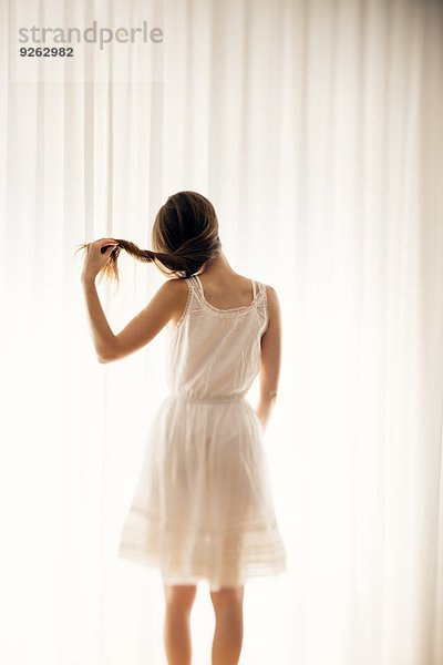 Junge Frau spielt mit ihrem Haar vor einem weißen Vorhang  Rückansicht
