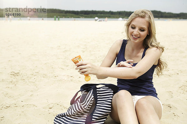 Lächelnde junge Frau am Strand sitzend mit Sonnencreme