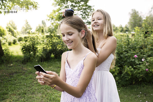Mädchen benutzt ihr Smartphone  während ihre Schwester ihr Haar bürstet.