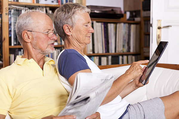 Seniorenpaar sitzt auf der Couch mit Zeitung mit digitalem Tablett
