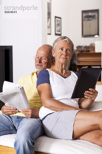 Seniorenpaar sitzt auf der Couch und liest Zeitung mit digitalem Tablett.
