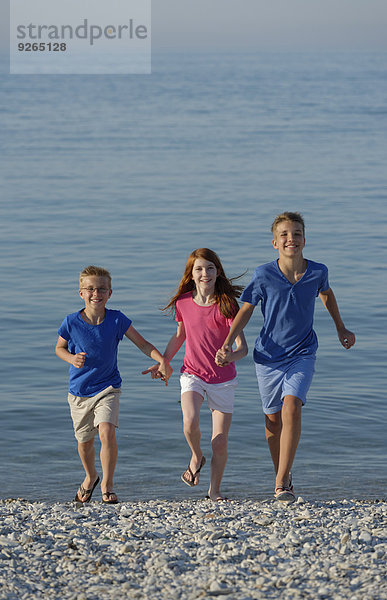 Italien  drei lächelnde Kinder laufen Hand in Hand am steinigen Strand vor dem Meer.