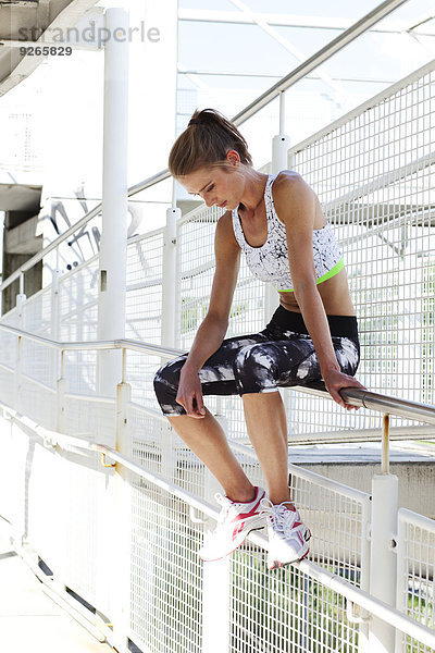 Junge Frau in Sportbekleidung auf einem Geländer einer Fußgängerbrücke sitzend