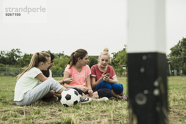 Vier weibliche Teenager-Freunde  die mit dem Smartphone auf dem Fußballplatz sitzen.