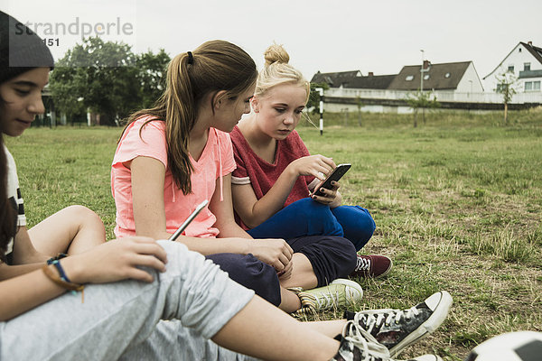 Vier weibliche Teenager-Freunde  die mit ihren Smartphones auf dem Fußballplatz sitzen.