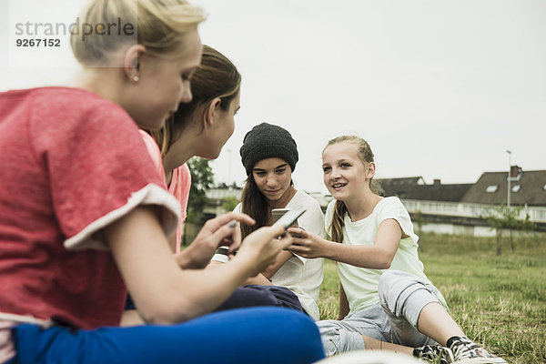 Vier weibliche Teenager-Freunde  die mit ihren Smartphones auf dem Fußballplatz sitzen.