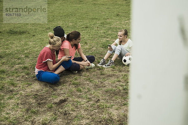 Vier weibliche Teenager-Freunde  die mit ihren Smartphones auf dem Fußballfeld sitzen.