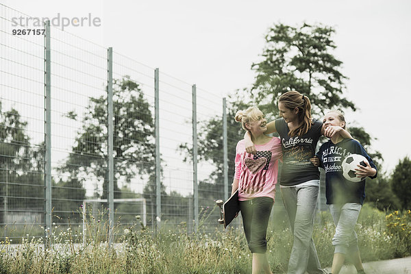 Drei fröhliche Teenagermädchen mit Fußball und Skateboard laufen nebeneinander.