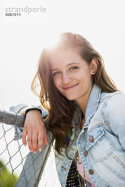Porträt eines lächelnden Teenagermädchens in Jeansjacke auf einem Zaun lehnend