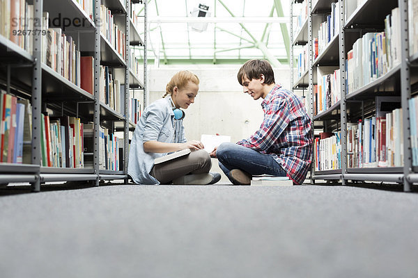 Zwei Studenten in einer Universitätsbibliothek sitzen auf dem Boden und lesen Bücher.