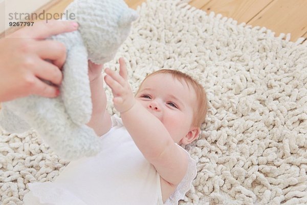 Weibliche Hand hält Teddybär für kleines Mädchen  das auf einem Teppich liegt