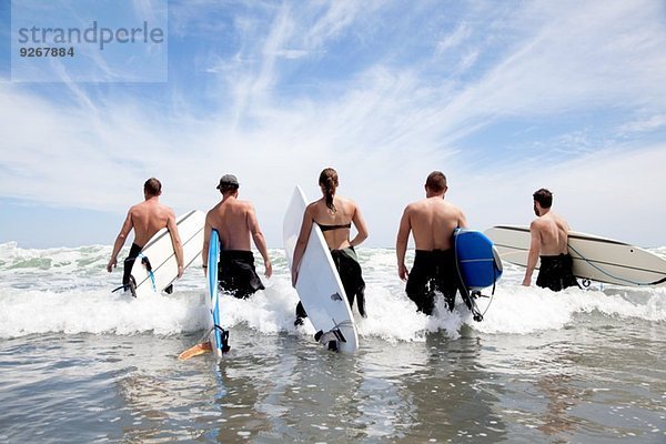 Rückansicht einer Gruppe männlicher und weiblicher Surferfreunde  die mit Surfbrettern ins Meer waten.