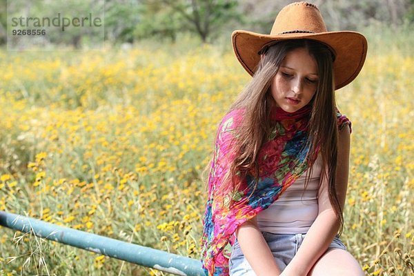 Mürrisches Mädchen mit Cowboyhut auf dem Zaun sitzend  das nach unten blickt.