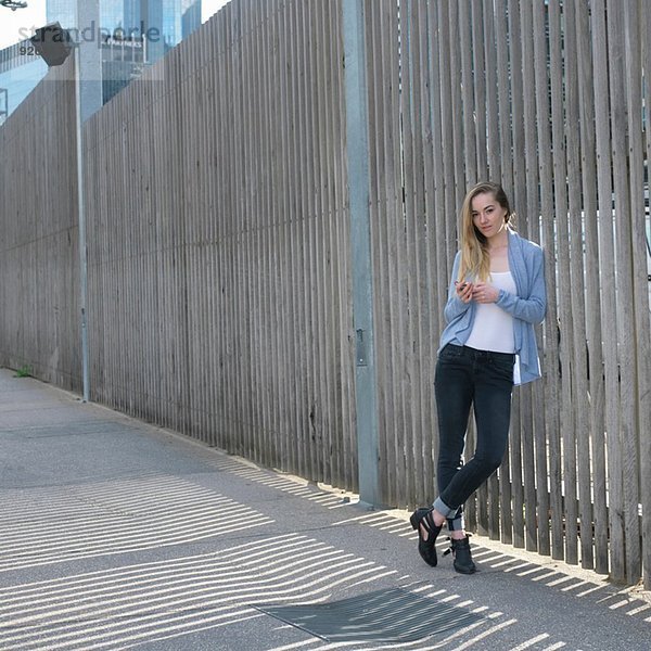 Porträt einer jungen Frau mit Smartphone am Zaun gelehnt