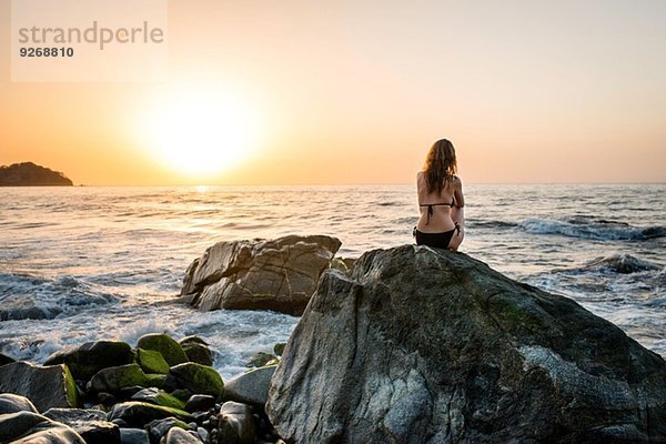 Junge Frau auf Felsen im Meer sitzend  Rückansicht