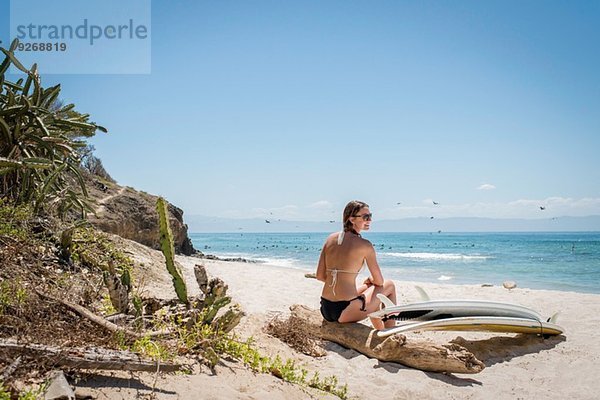 Junge Frau auf Treibholz am Strand sitzend  Surfbrett an ihrer Seite