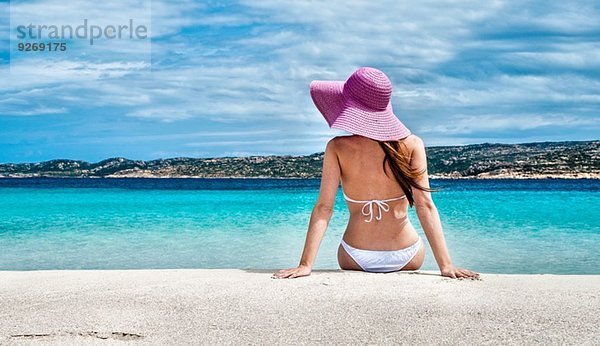 Rückansicht der jungen Frau mit Bikini und Sonnenhut am Strand  La Maddalena  Sardinien  Italien