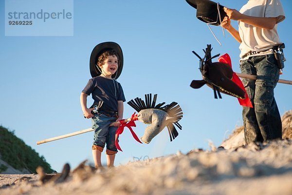 Zwei Brüder verkleidet als Cowboys mit Hobbypferden im Sand