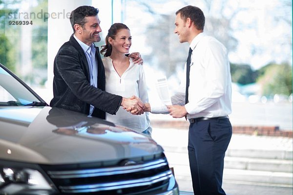 Mittleres erwachsenes Paar schüttelt sich die Hand mit dem Verkäufer im Autohaus.