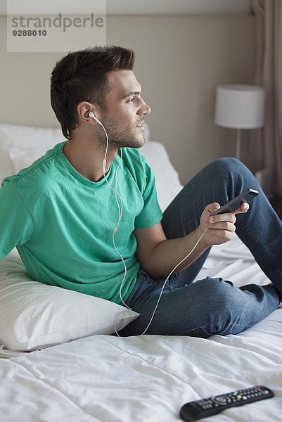 Junger Mann sitzt auf dem Bett und hört Musik mit dem Smartphone.