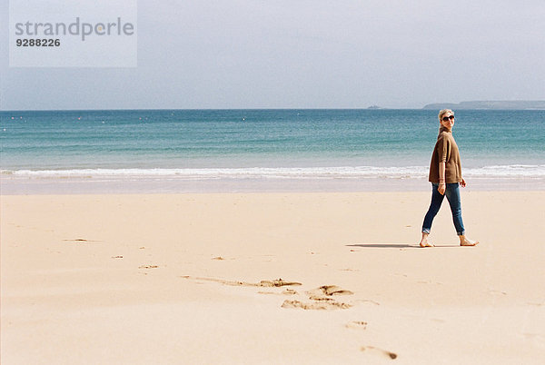 Eine Frau  die barfuß am Strand spazieren geht und dabei Fußspuren im Sand hinterlässt.