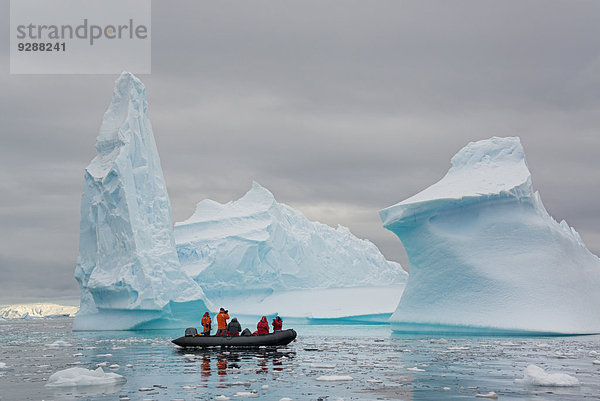 Menschen in kleinen aufblasbaren Zodiac-Rib-Booten  die auf dem ruhigen Wasser um kleine Inseln der Antarktischen Halbinsel an hoch aufragenden  gemeißelten Eisbergen vorbeifahren.