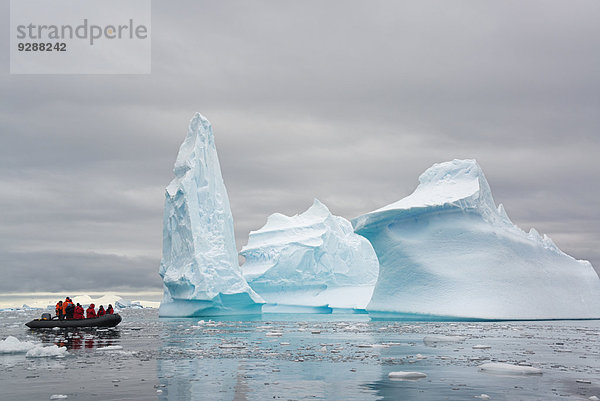 Menschen in kleinen aufblasbaren Zodiac-Rib-Booten  die auf dem ruhigen Wasser um kleine Inseln der Antarktischen Halbinsel an hoch aufragenden  gemeißelten Eisbergen vorbeifahren.