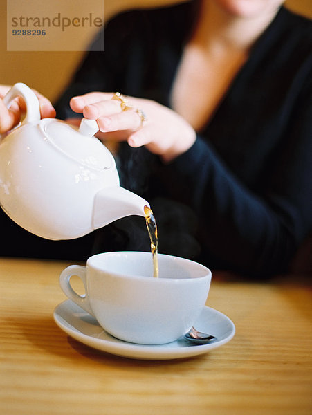 Eine sitzende Frau gießt aus einer Teekanne aus weißem Porzellan eine Tasse Tee aus.