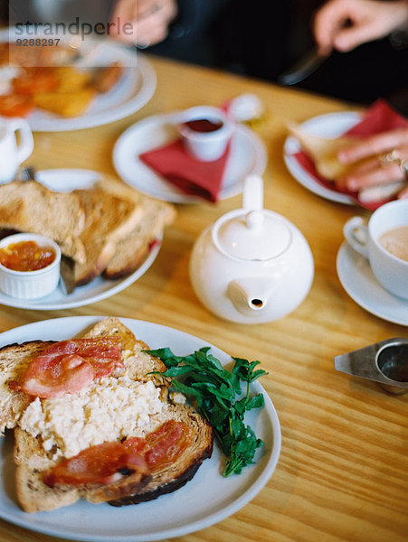 Ein Tisch  der mit Essenstellern gedeckt ist. Ein gekochtes Frühstück mit Speck  Eiern und Toast und einer Kanne Tee. Zwei sitzende Personen