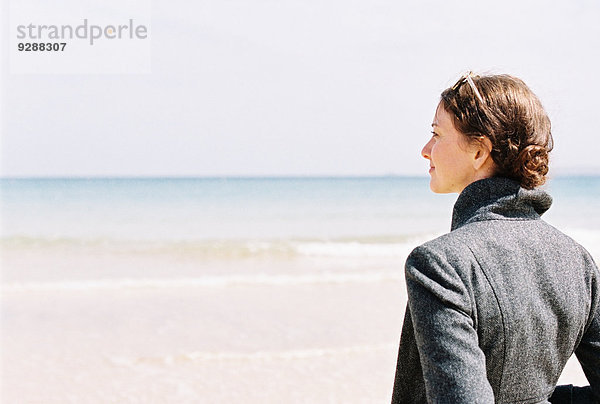 Eine Frau in einer grauen Jacke mit Blick auf den Strand und das Meer.